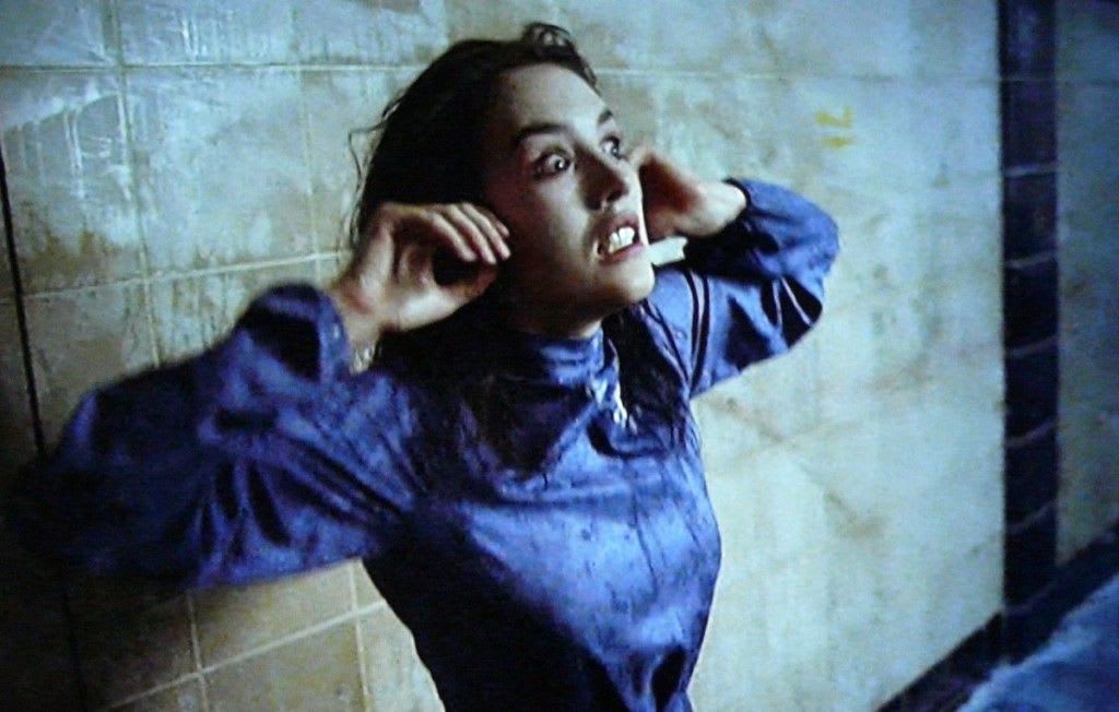 Screenshot of Anna in Possession - white woman wearing a blue dress stands before a grimy tile wall. Her arms are raised, hands beside her face, her expression is one of horrific strain.