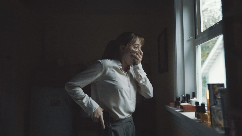 Screenshot from Longlegs featuring a white actress in a white shirt, hand over her face in shock as she look out a window.